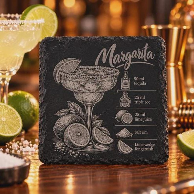 A square black slate coaster is engraved with a Margarita illustration, recipe, and title, set against a blurred bar background with limes and a cocktail glass.