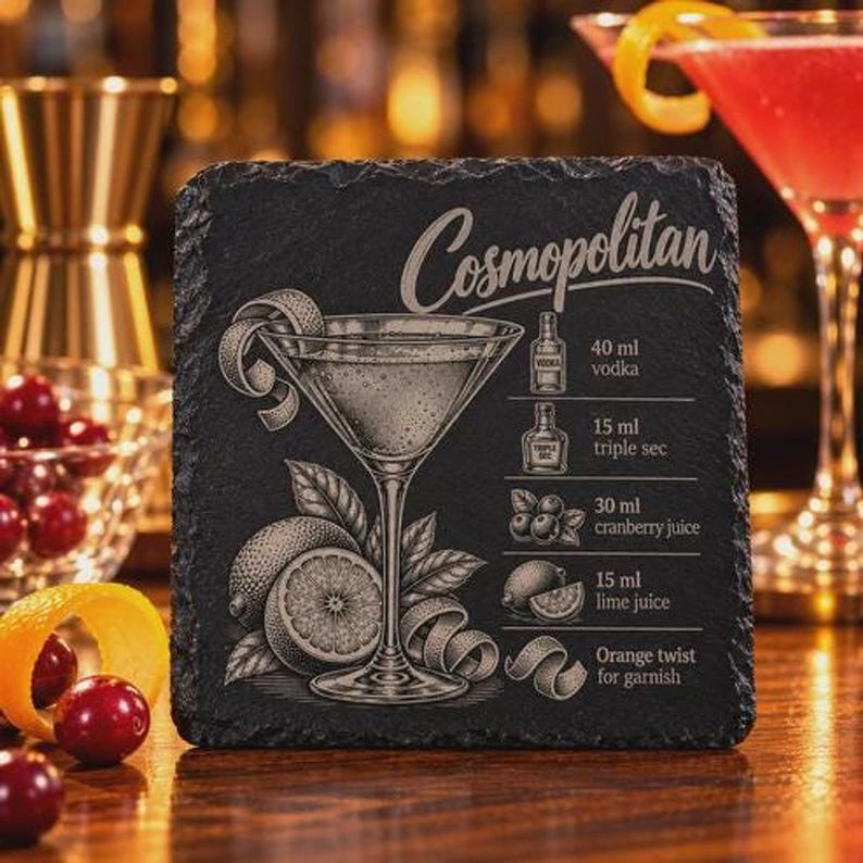 A square black slate coaster is engraved with a Cosmopolitan illustration, recipe, and title, displayed on a wooden surface near a red cocktail glass.