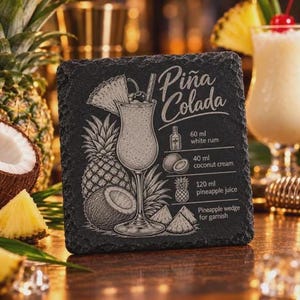 A square black slate coaster features an engraved Pina Colada design with a pineapple illustration, recipe, and title on a wooden bar surface.