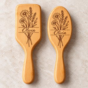 May include: Two light brown wooden hairbrushes with a smooth finish. Each brush features a dark brown laser-engraved illustration of a wildflower bouquet and the name Olivia centered within a horizontal line design on the handle.
