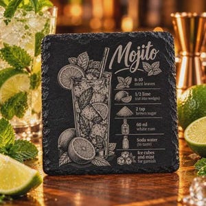 A square black slate coaster features an engraved Mojito illustration, recipe, and title, displayed on a wooden surface with mint leaves and lime wedges.