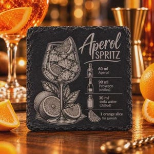 A square black slate coaster is engraved with an Aperol Spritz cocktail illustration, recipe, and title, placed on a wooden bar next to an orange slice.