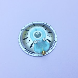 May include: A circular silver-coloured metal component featuring a series of small ventilation holes arranged in a radial pattern. A translucent light blue silicone piece is attached to the centre, secured by two screws, with a small brass connector.