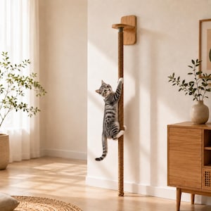 Puede incluir: Un rascador para gatos montado en la pared que presenta un cilindro vertical alto envuelto en una cuerda de sisal gruesa de color natural. El poste está asegurado a la pared con un soporte de madera de tono claro en la parte superior.