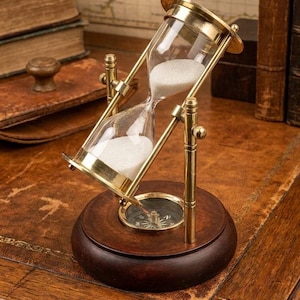 May include: A brass hourglass with white sand, mounted on a circular dark wood base that features an integrated magnetic compass. The hourglass is held by a rotating brass frame with vertical supports, allowing the glass bulb to tilt.