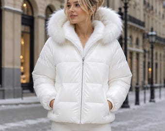 May include: A white puffer jacket with a glossy finish, featuring a large hood trimmed with thick white faux fur. The jacket has a full-length silver zipper, long sleeves with elastic cuffs, and two front pockets, providing a clean, bright look.