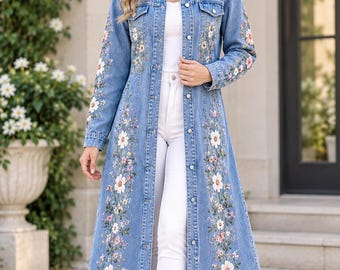 May include: A long-length blue denim duster coat featuring a classic collar, button-down front, and long sleeves. The garment is decorated with intricate floral embroidery in shades of white, pink, and green along the front panels and sleeves.