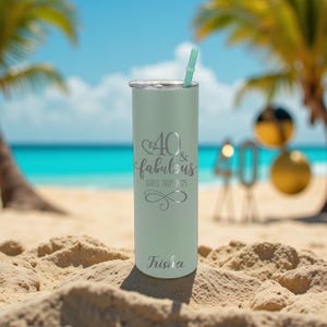 Puede incluir: Un vaso t&eacute;rmico verde menta con tapa y pajita plateadas, sobre arena. El vaso est&aacute; inscrito con "40 & fabulous GIRLS TRIP 2025" y el nombre "Trisha". El fondo presenta una escena de playa con palmeras y el oc&eacute;ano.