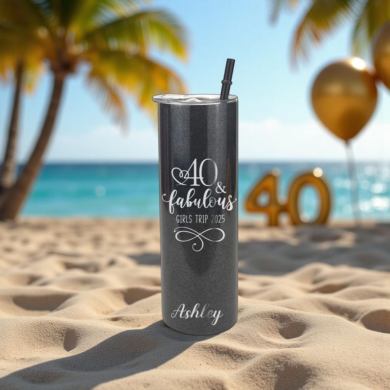 Puede incluir: Un vaso de color gris oscuro con una pajita negra, personalizado con el nombre "Ashley". El vaso presenta el texto "40 & fabulous GIRLS TRIP 2025" en escritura blanca. El fondo incluye una escena de playa con palmeras y globos dorados.