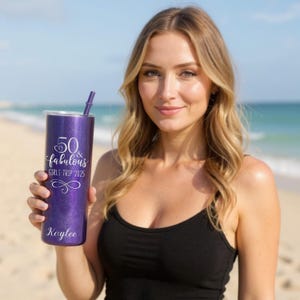 K&ouml;nnte beinhalten: Ein lila Glitzerbecher mit Strohhalm, mit dem Text "50 & Fabulous GIRLS TRIP 2025" und dem Namen "Kaylee". Der Becher ist zylindrisch und hat einen silbernen Deckel. Der Hintergrund ist ein Strand mit dem Meer.