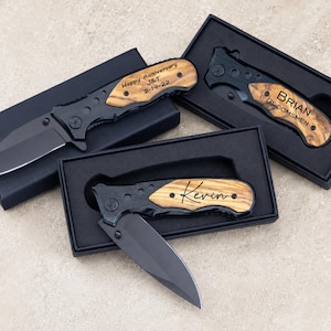 May include: Three folding pocket knives with matte black blades and handles featuring wood grain inlays. Each knife is housed in a black gift box. The knives are engraved with the text Happy Anniversary J&T 2-14-22, Brian Groomsmen, and Kevin.