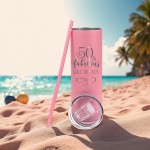 K&ouml;nnte beinhalten: Ein rosa isolierter Becher mit einem passenden rosa Strohhalm. Auf dem Becher steht in silberner Schrift der Text "50 & fabulous GIRLS TRIP 2025". Der Deckel ist transparent und der Becher steht an einem Sandstrand mit dem Meer im Hintergrund.