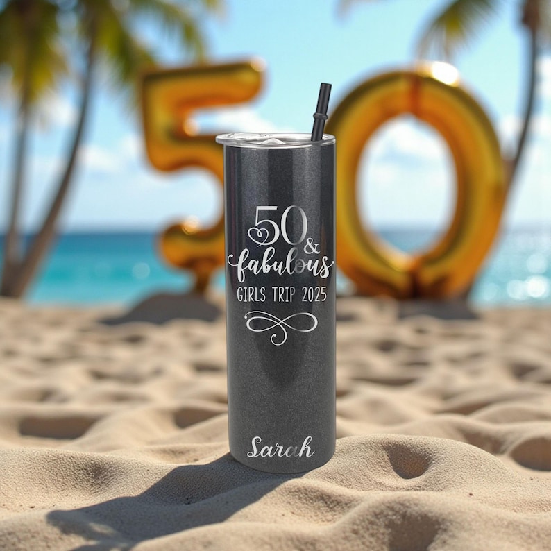 K&ouml;nnte beinhalten: Ein dunkelgrauer Becher mit einem schwarzen Strohhalm, mit dem Text "50 & fabulous GIRLS TRIP 2025" in wei&szlig;er Schrift. Der Name "Sarah" ist ebenfalls auf dem Becher aufgedruckt. Goldene Zahlenballons sind im Hintergrund.