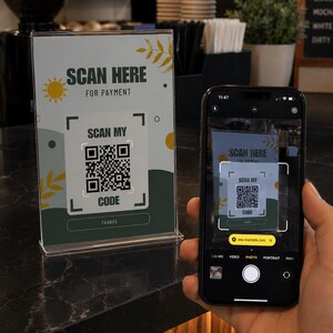 Könnte beinhalten: Ein transparenter Acryl-Schilderständer mit einer weißen Karte, die den Text SCAN HERE FOR PAYMENT und SCAN MY CODE über einem QR-Code zeigt. Ein Smartphone wird davor gehalten und nutzt die Kamera-App, um den angezeigten QR-Code zu scannen.