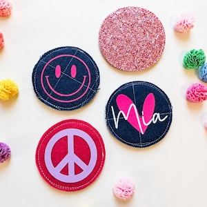 Könnte beinhalten: Vier runde Untersetzer, darunter einer mit rosa Glitzer, einer in Dunkelblau mit rosa Smiley, einer in Dunkelblau mit rosa Herz und dem Namen Mia in weißer Schreibschrift sowie einer in Rot mit hellviolettem Friedenszeichen.