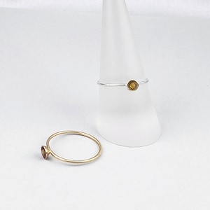 May include: Two simple gold rings with a small round gemstone in the center. The rings are on a white cone-shaped display stand.