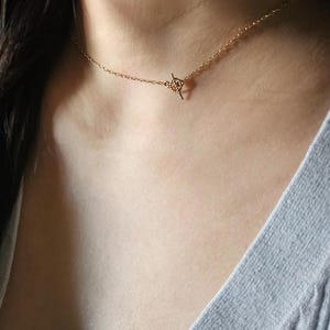 以下が含まれることがあります： 繊細なゴールドカラーのネックレス、トグルクラスプ付き。 ネックレスは首にかけられ、明るいグレーのトップスに映えます。 チェーンは細く、クラスプは小さな装飾要素です。