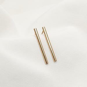 Puede incluir: Un par de pendientes de barra dorados. Los pendientes son rectos, delgados y tienen un diseño minimalista. Se encuentran sobre un fondo de tela blanca texturizada.