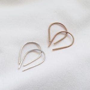 May include: Two pairs of earrings, one gold and one silver, are displayed on a white surface. The earrings have a looped design with a twisted rope texture. The gold earrings are on the right, and the silver earrings are on the left.