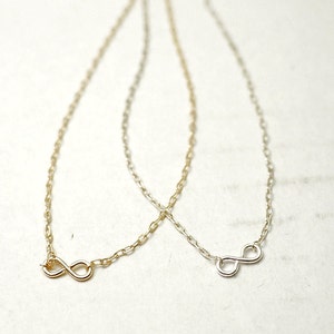 Könnte beinhalten: Zwei Halsketten mit einem Unendlichkeitszeichen, eine in Gold und eine in Silber, auf weißem Hintergrund.