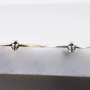 Puede incluir: Dos anillos delicados con forma de calavera, uno en oro y otro en plata, sobre un fondo blanco.