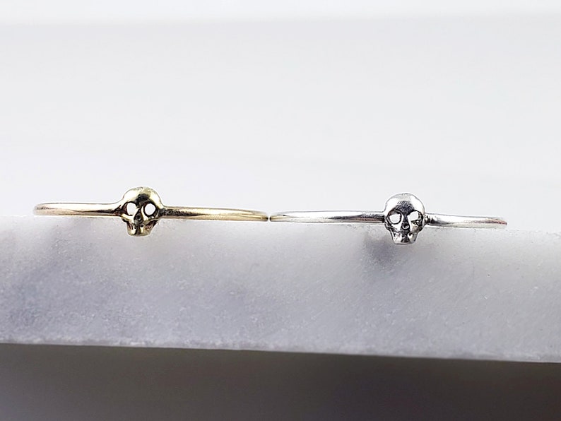 Puede incluir: Dos anillos delicados con forma de calavera, uno en oro y otro en plata, sobre un fondo blanco.