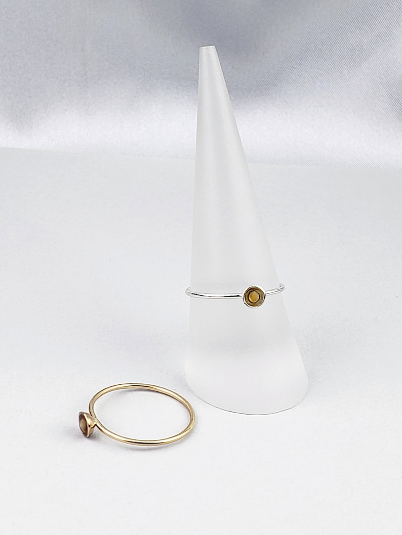 May include: Two simple gold rings with a small round gemstone in the center. The rings are on a white cone-shaped display stand.
