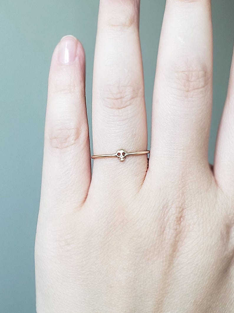 Puede incluir: Un delicado anillo de oro con un peque&ntilde;o dise&ntilde;o de calavera. El anillo se lleva en el dedo anular de una mano.