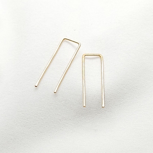 May include: Two gold-colored hair pins in a U-shape are displayed on a white, textured surface. One pin is slightly larger than the other. The pins are minimalist in design and appear to be made of metal.