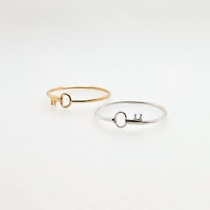 May include: Two simple key-shaped rings, one gold and one silver. The rings are delicate and minimalist.