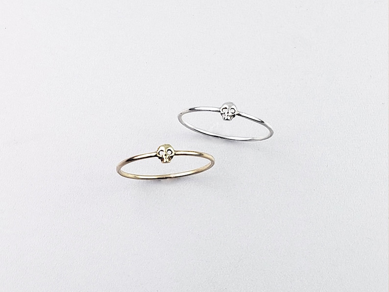 Puede incluir: Dos anillos simples y delicados con un peque&ntilde;o dise&ntilde;o de calavera. El anillo de la izquierda es de oro y el anillo de la derecha es de plata.