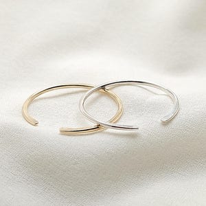 Puede incluir: Dos brazaletes minimalistas de extremo abierto. Uno es dorado, el otro plateado. Los brazaletes descansan sobre una tela blanca texturizada. El diseño es simple y elegante.