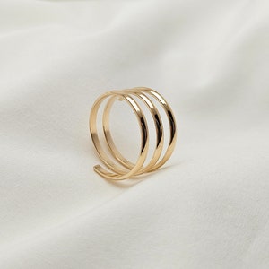 May include: A gold-colored ring with a spiral design. The ring features four parallel bands that curve around, creating a unique and modern look. The ring is set against a white background.