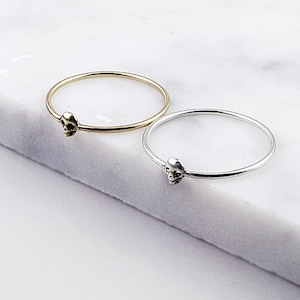 Puede incluir: Dos anillos simples de plata con un peque&ntilde;o encanto de calavera en cada uno. El anillo de la izquierda es de oro y el anillo de la derecha es de plata.