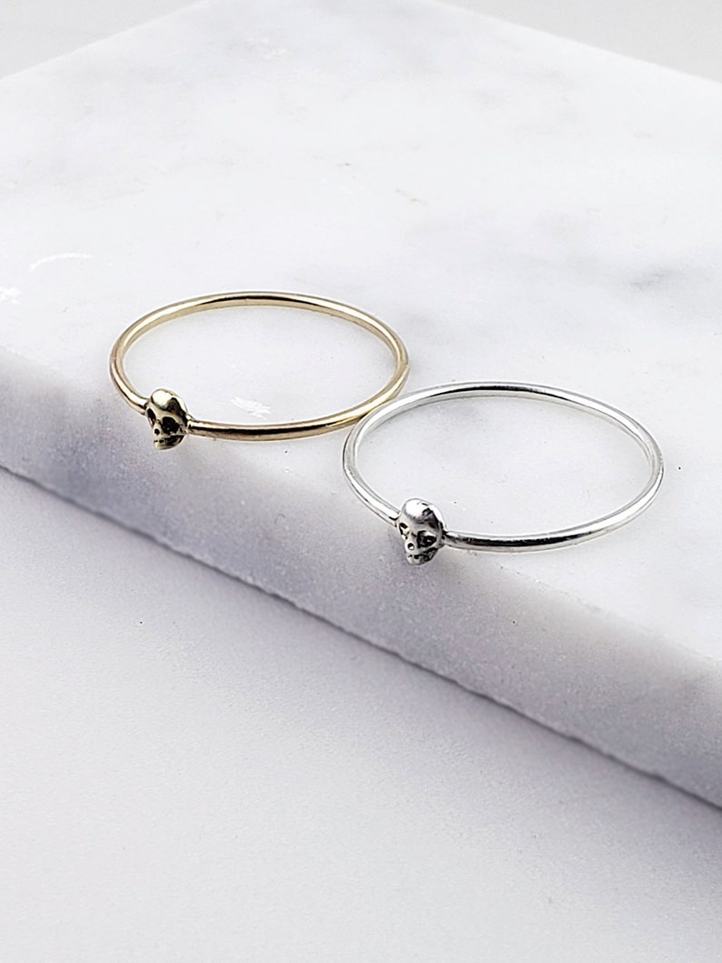 Puede incluir: Dos anillos simples de plata con un peque&ntilde;o encanto de calavera en cada uno. El anillo de la izquierda es de oro y el anillo de la derecha es de plata.