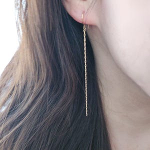 Puede incluir: Primer plano de un pendiente de cadena dorada. El pendiente es una cadena larga y delicada que cuelga de la oreja. La cadena está hecha de pequeños eslabones. El pendiente es una joya sencilla y elegante.