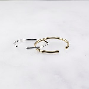 May include: Two nose rings, one silver and one gold, are shown on a white surface. The rings are open and have a simple, rounded design.