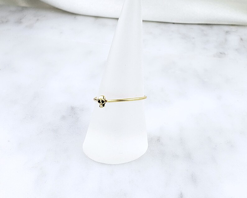 Pode incluir: Um anel de ouro com um pequeno design de caveira. O anel &eacute; exibido em um suporte c&ocirc;nico branco.