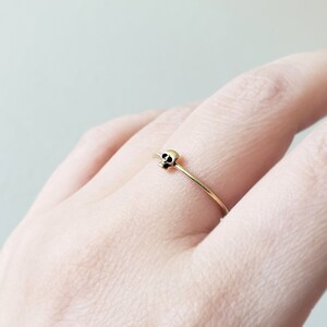 Pode incluir: Um anel de ouro com um pequeno design de caveira. O anel &eacute; usado no dedo de uma pessoa.