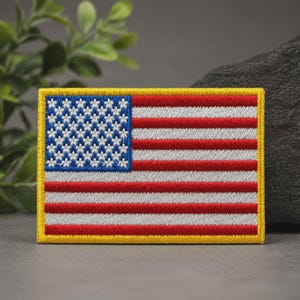 May include: A rectangular embroidered patch featuring the United States flag design. The canton is blue with white star-shaped embroidery, and the field consists of alternating horizontal red and white stripes, all bordered by a thick yellow frame.