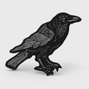 May include: An embroidered patch in the shape of a raven, featuring detailed black and dark gray stitching that defines the bird's feathers, beak, and talons. The design captures the bird in a side profile, standing with its wings folded.