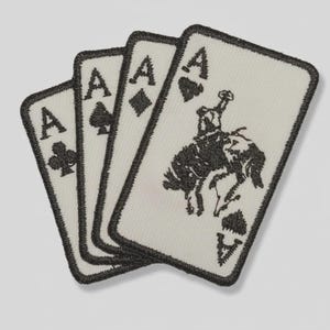 May include: A set of four rectangular embroidered patches featuring playing card designs. Each patch has a white background with black stitched borders and details, including the Ace of Clubs, Ace of Spades, Ace of Diamonds, and an Ace of Spades with a cowboy riding a bucking horse.