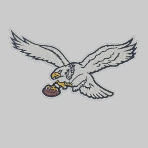 May include: An embroidered patch featuring a white eagle with dark outlines, a yellow beak, and yellow talons clutching a brown football. The eagle is depicted in flight with wings fully extended, showcasing detailed stitching and a textured appearance.