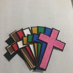 May include: A collection of embroidered cross patches arranged in a fan shape. The patches feature a black border and come in various solid colors including pink, blue, green, gold, white, red, brown, and black, each with a textured thread finish.