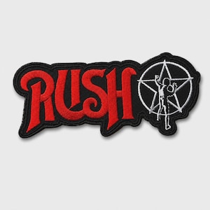 May include: A black embroidered patch featuring the red text RUSH in a stylized font, followed by a white circular emblem containing a five-pointed star and a silhouette of a person with arms raised, all outlined with a thick black border.