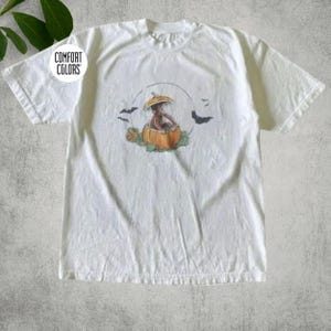 Puede incluir: Una camiseta blanca de manga corta con un gráfico de un mono marrón sentado dentro de una calabaza naranja. El diseño incluye un contorno circular fino, hojas verdes y murciélagos negros. El texto COMFORT COLORS está impreso en un círculo.