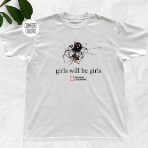 Könnte beinhalten: Ein weißes Kurzarm-T-Shirt mit einer Grafik einer Schwarzen Witwe auf einem Spinnennetz. Unter der Spinne steht der Text "girls will be girls" in Schwarz, gefolgt vom National Geographic-Logo in einem roten Quadrat mit schwarzem Text.