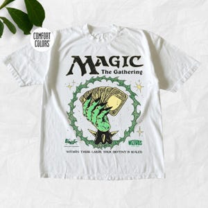 May include: A white short-sleeve t-shirt featuring a graphic of a green hand holding playing cards inside a green circular border. The text reads Magic The Gathering, Wizards, and Within these cards, your destiny is sealed. A Comfort Colors logo is visible.