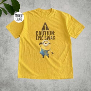 May include: A yellow short-sleeve t-shirt featuring a graphic of a single-eyed character wearing blue overalls. Above the character is a black triangle with an exclamation point and the text CAUTION: EPIC SWAG. A Comfort Colors logo is visible.