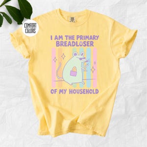 Könnte beinhalten: Ein blassgelbes T-Shirt mit einer Grafik einer hellgrünen Maus mit Sonnenbrille, die ein Baguette und ein Martiniglas hält. Der Text lautet I AM THE PRIMARY BREADLOSER OF MY HOUSEHOLD, platziert vor vertikalen Pastellstreifen und Sternakzenten.